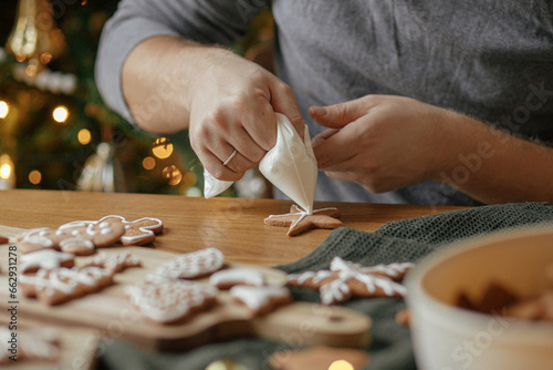 Decorating gingerbread cookies with icing on rustic wooden table at christmas tree golden lights. Atmospheric Christmas holiday traditions. Man decorating cookies with sugar frosting photo