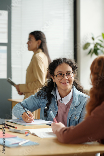 Cute smiling schoolgirl with pen and copybook looking at classmate at lesson of English language during discussion of new grammar rules