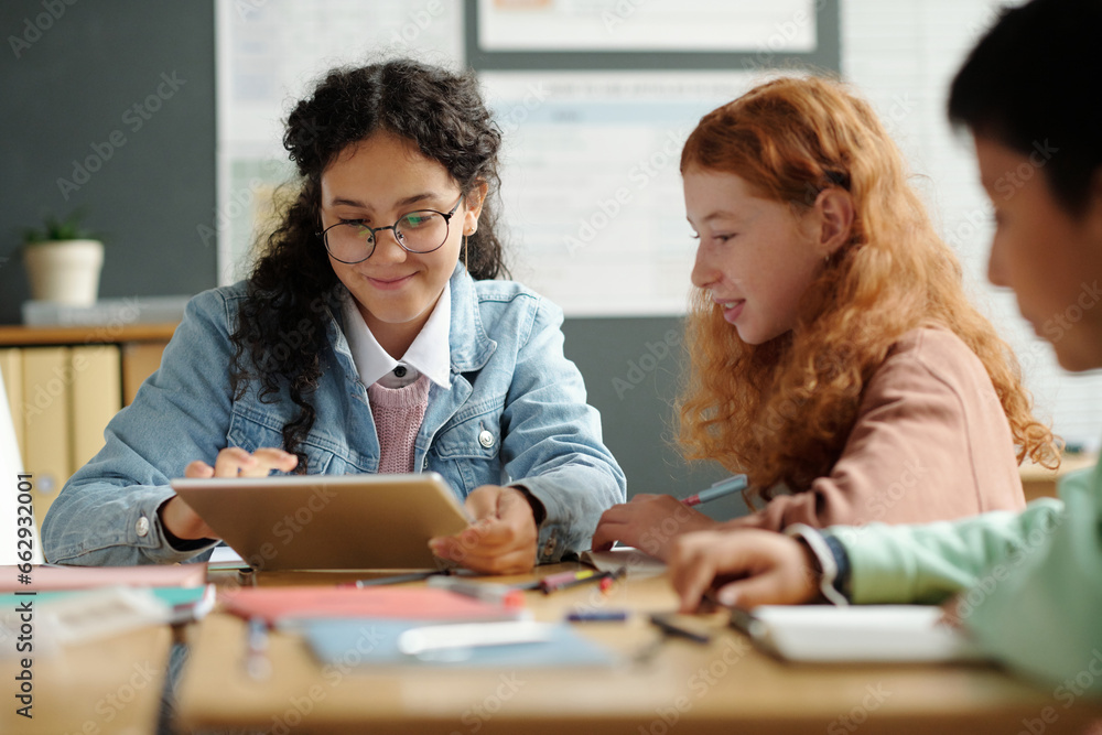 Fototapeta premium Two cute intercultural schoolgirls preparing presentation of English words or new grammar rules while one of them touching gadget screen