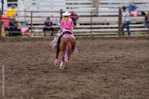 A cowgirl is riding a horse at a rodeo in a dirt arena. The horse is brown and has sequence on its back . She has a pink blouse and a white hat. 
