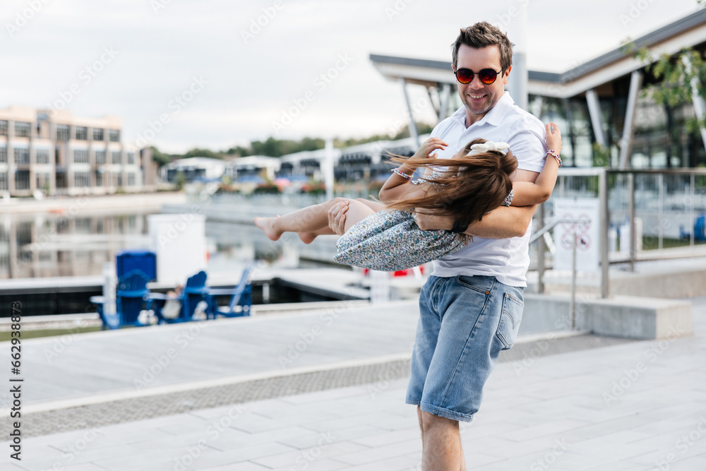 Fototapeta premium A happy father holds his daughter in his arms and spins her around. Dad and daughter are having fun walking outside. Happy fatherhood and childhood. Dad and his little daughter having fun