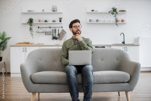 Handsome caucasian man in eyeglasses sitting on couch with wireless laptop and thoughtfully looking aside. Bearded young guy reflecting and thinking at home during distance work.