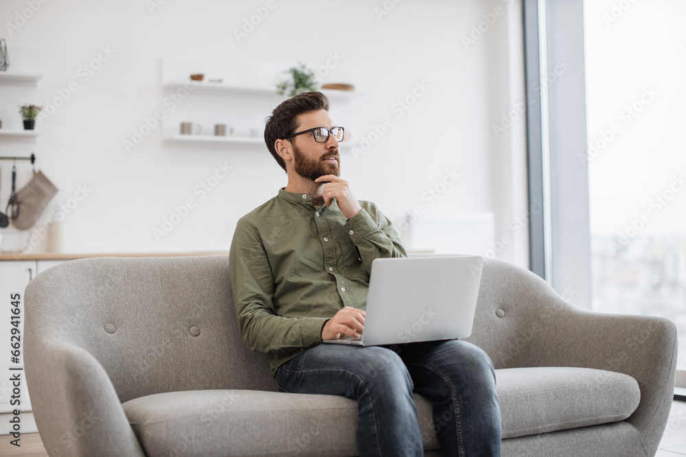 Handsome caucasian man in eyeglasses sitting on couch with wireless laptop and thoughtfully looking aside. Bearded young guy reflecting and thinking at home during distance work.