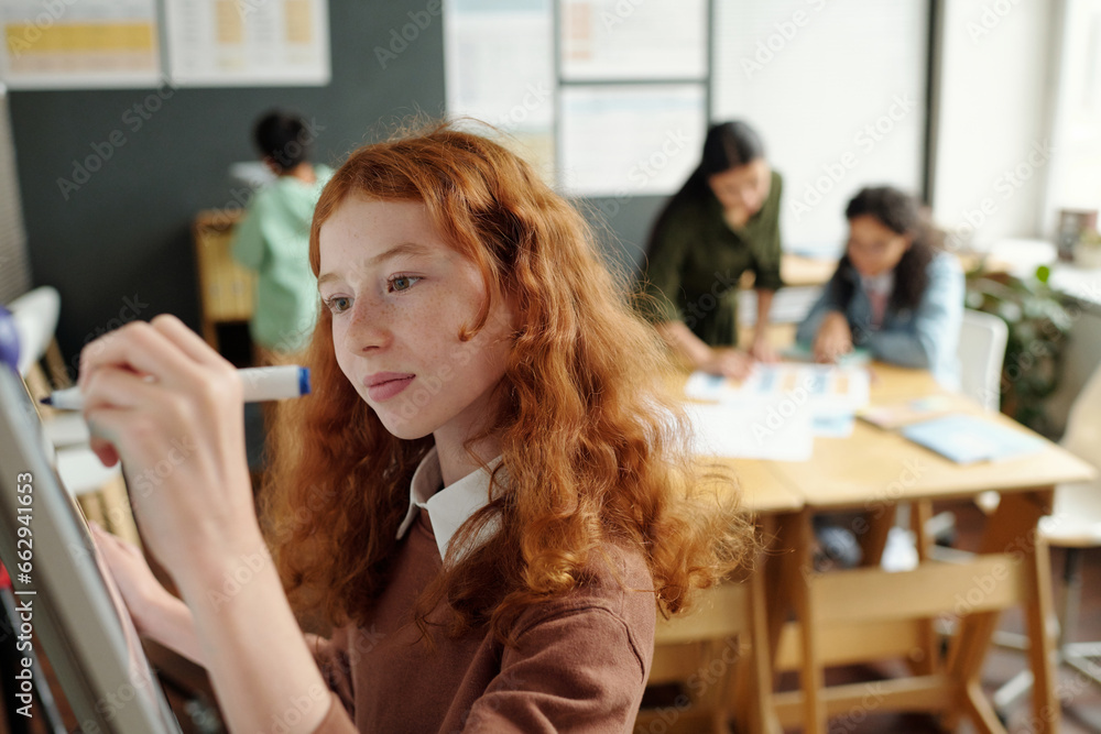 Focus on cute schoolgirl writing grammar rules on whiteboard at lesson while preparing for presentation against classmates and teacher Focus on cute schoolgirl writing grammar rules on whiteboard at lesson while preparing for presentation against classmates and teacher