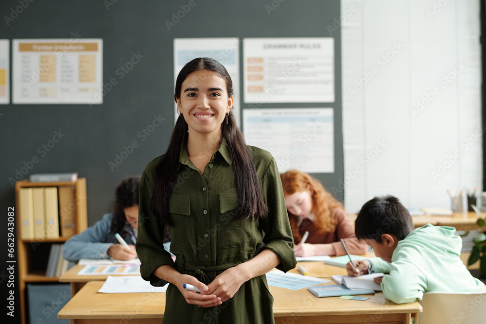 Fototapeta premium Young professional teacher of English language looking at camera while standing against group of intercultural schoolkids making notes