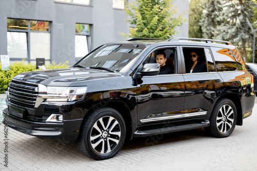 Side view of a luxury SUV vehicle with male driver and business lady on a backseat. Concept of luxury transportation and personal driver