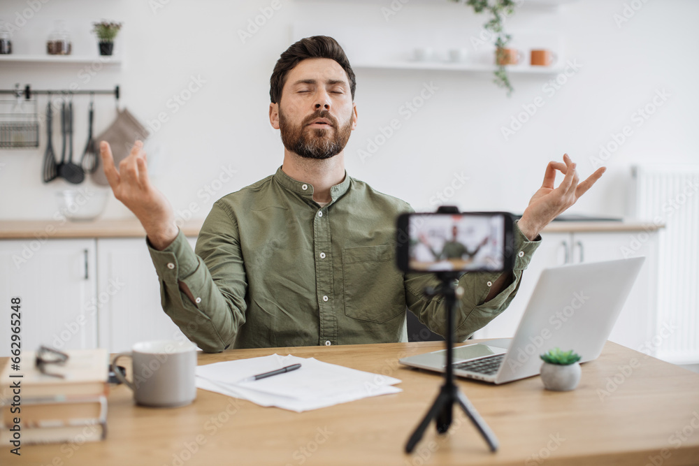 Concentrated bearded vlogger sitting in front of smartphone fixed on tripod and live streaming at home. Calm man holding hands in mudra gesture and shooting tutorial of yoga meditation. Concentrated bearded vlogger sitting in front of smartphone fixed on tripod and live streaming at home. Calm man holding hands in mudra gesture and shooting tutorial of yoga meditation.