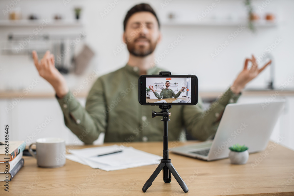 Blurred background of male streamer meditating with closed eyes and holding mudra gesture while filming online course at home. Focus on wireless smartphone fixed on tripod and recording video. Blurred background of male streamer meditating with closed eyes and holding mudra gesture while filming online course at home. Focus on wireless smartphone fixed on tripod and recording video.