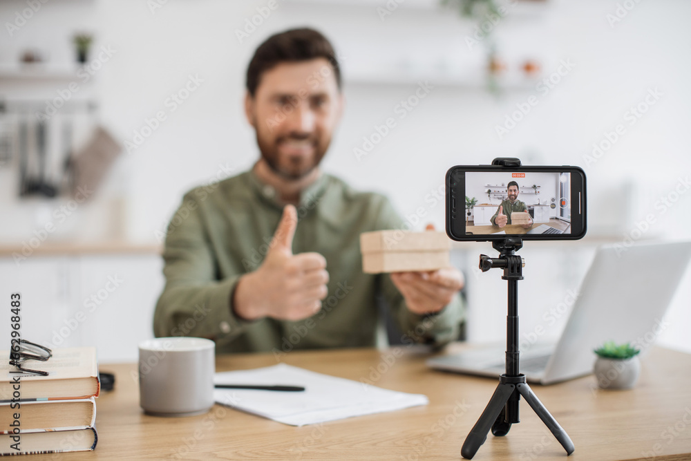 Modern smartphone on tripod recording video of joyful caucasian influencer making unpacking of cardboard parcel at home showing thumb up Modern smartphone on tripod recording video of joyful caucasian influencer making unpacking of cardboard parcel at home showing thumb up