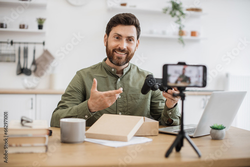 Handsome professional blogger showing impression while demonstrating new joystick to subscribers by cellphone camera. Bearded man creating new video content to social media at home. © sofiko14
