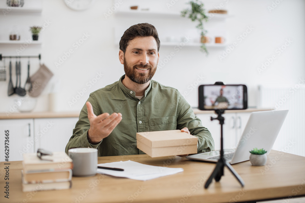 Caucasian young vlogger recording new video on smartphone about unboxing modern technologies. Bearded male person holding box and enjoying reviewing online purchase. Concept of working at home. Caucasian young vlogger recording new video on smartphone about unboxing modern technologies. Bearded male person holding box and enjoying reviewing online purchase. Concept of working at home.