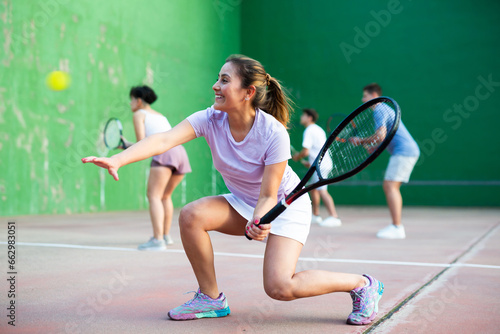 Active fit young hispanic woman playing frontenis on open court on summer day, hitting ball with strung tennis racquet to score to opposing team. Popular Spanish sports.. © JackF