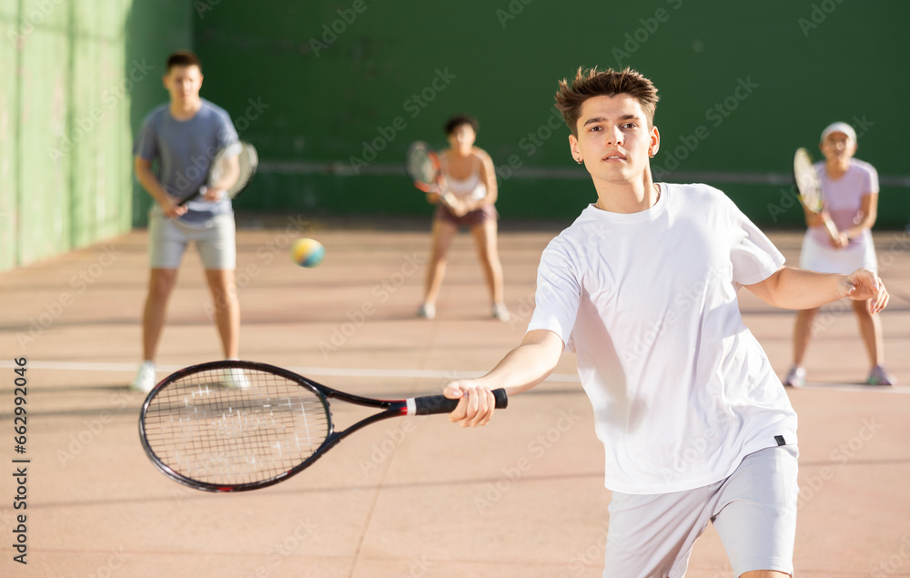 Concentrated young Latin American frontenis player swinging string racquet to hit ball on outdoor walled court on sunny summer day. Sport and active lifestyle concept