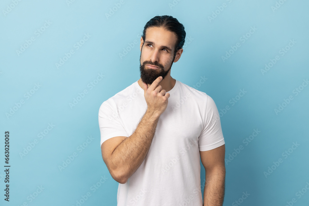Portrait of thoughtful handsome man with beard wearing white T-shirt holding his chin and pondering idea, confused not sure about solution. Indoor studio shot isolated on blue background. Portrait of thoughtful handsome man with beard wearing white T-shirt holding his chin and pondering idea, confused not sure about solution. Indoor studio shot isolated on blue background.