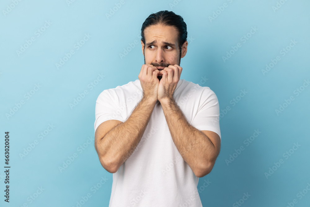 Troubles and worries. Portrait of man with beard wearing white T-shirt biting nails, terrified about problems, suffering phobia, anxiety disorder. Indoor studio shot isolated on blue background.