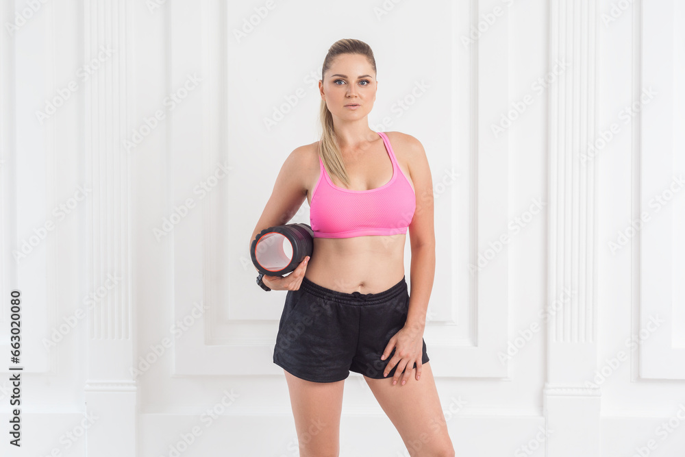 Portrait of confident athletic sporty beautiful blonde woman with perfect body in pink top and black shorts standing, holding foam roll and looking at camera. Indoor studio shot.