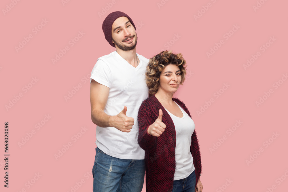 Portrait of cheerful young adult couple of friends standing together looking at camera and showing like gesture, thumbs up, agree, recommend. Indoor studio shot isolated on pink background. Portrait of cheerful young adult couple of friends standing together looking at camera and showing like gesture, thumbs up, agree, recommend. Indoor studio shot isolated on pink background.