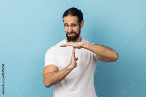 Portrait of bossy serious man with beard wearing white T-shirt showing time out gesture, looking with strict expression, deadline. Indoor studio shot isolated on blue background.
