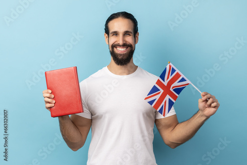 Portrait of smiling positive man with beard wearing white T-shirt holding book and Great Britain flag, education, learning English language. Indoor studio shot isolated on blue background. photo