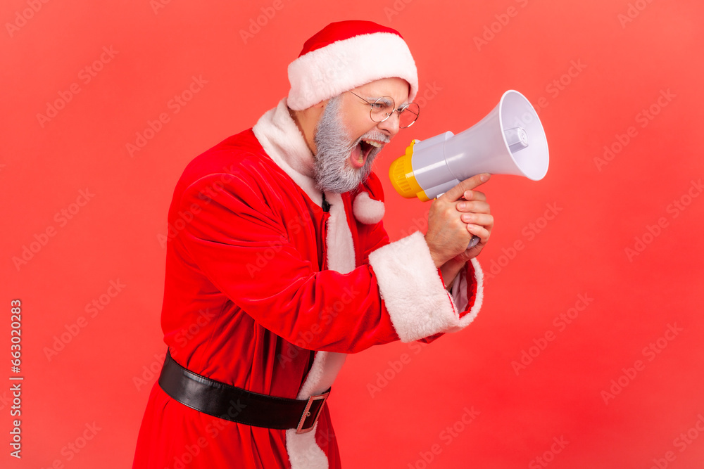 Side view of angry elderly man with gray beard wearing santa claus costume standing with megaphone in hands, shouting announcement, serious expression. Indoor studio shot isolated on red background. Side view of angry elderly man with gray beard wearing santa claus costume standing with megaphone in hands, shouting announcement, serious expression. Indoor studio shot isolated on red background.