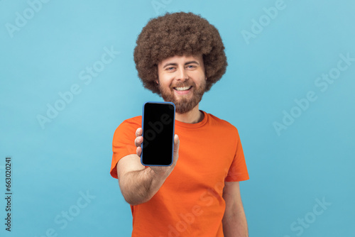 Portrait of delighted man with Afro hairstyle wearing orange T-shirt showing smart phone with blank black screen, presenting area for advertisement. Indoor studio shot isolated on blue background.