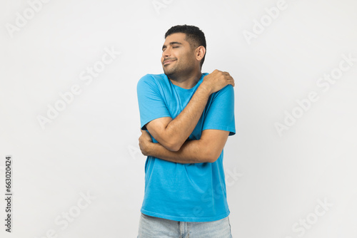 Portrait of happy optimistic smiling unshaven man wearing blue T- shirt standing hugging himself, looks satisfied, expressing happiness. Indoor studio shot isolated on gray background.