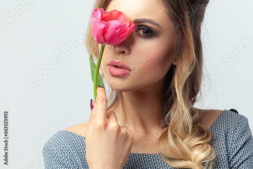 Portrait of adorable mysterious blonde woman fashion model with makeup and hairstyle, hiding her face behind tulip, looking at camera. Indoor studio shot isolated on gray background.