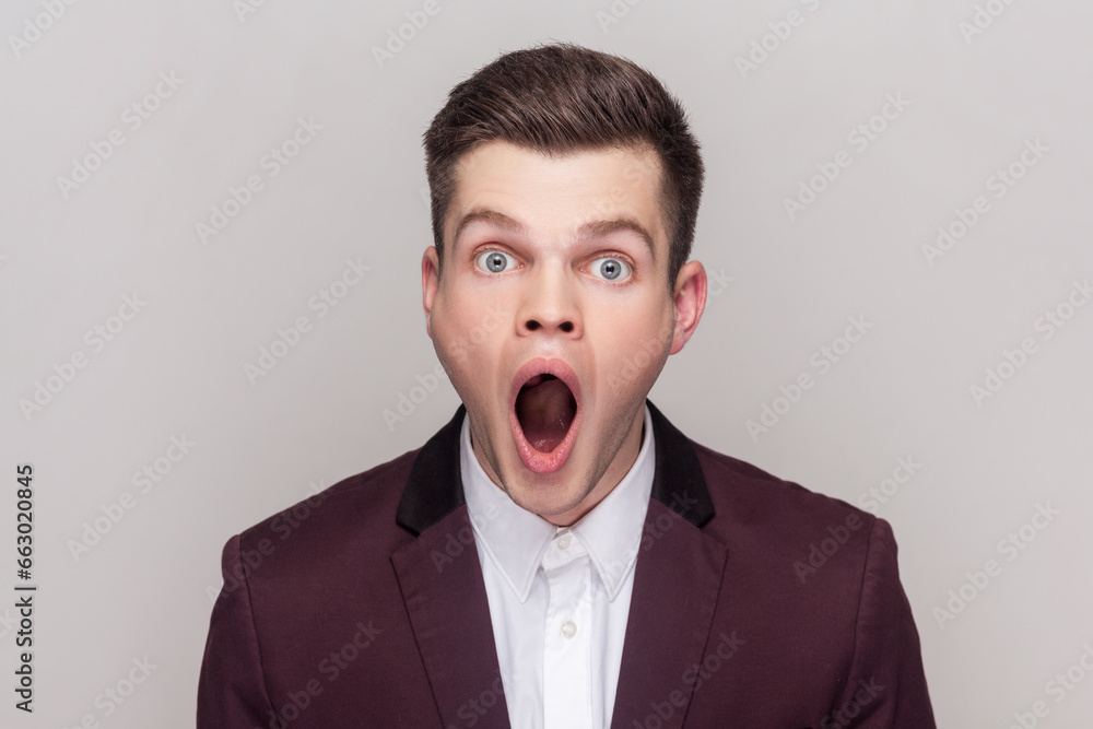 Shocked surprised handsome young man looking at camera with big eyes and open mouth sees something astonished, wearing violet suit and white shirt. Indoor studio shot isolated on grey background. Shocked surprised handsome young man looking at camera with big eyes and open mouth sees something astonished, wearing violet suit and white shirt. Indoor studio shot isolated on grey background.