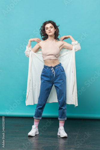Full length of beautiful woman in casual style attire standing looking at camera with serious face, keeps hands on her shoulders. Indoor studio shot isolated on blue background.