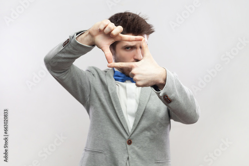 Portrait of focused concentrated bearded man standing looking at camera through frame from fingers, making photo, wearing grey suit and blue bow tie. Indoor studio shot isolated on gray background.