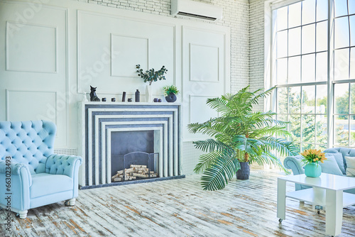 Fototapeta Naklejka Na Ścianę i Meble -  Beautiful apartment interior with large floor-to-ceiling windows in blue tones. The room contains pots with live plants, a sofa, a coffee table, an armchair, a decorative fireplace, and a white floor.