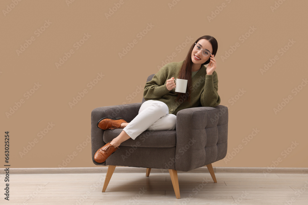 Beautiful woman with cup of drink sitting in armchair near beige wall indoors Beautiful woman with cup of drink sitting in armchair near beige wall indoors