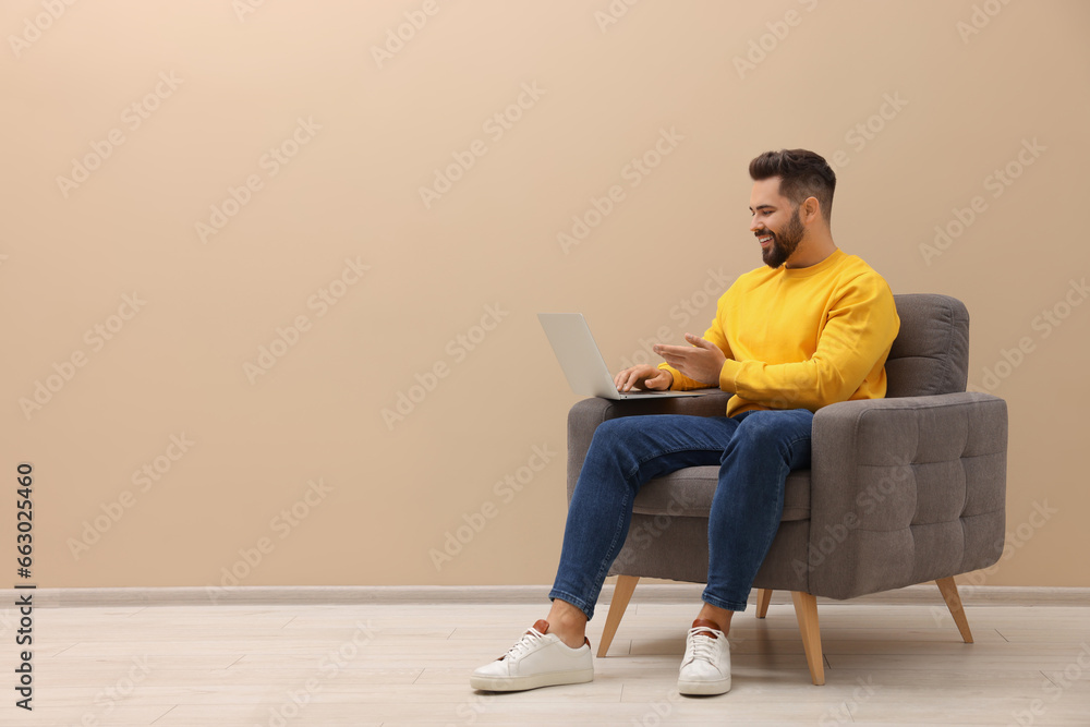 Handsome man with laptop sitting in armchair near beige wall indoors, space for text