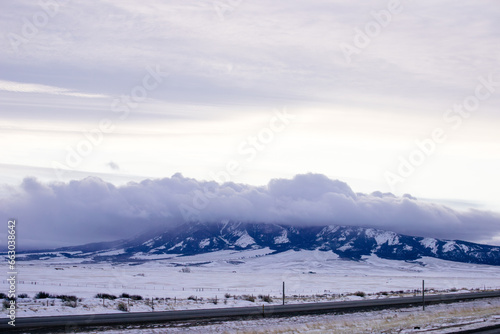 Beautiful winter landscape with a highway between blue snowy mountains, fluffy clouds on a sunset orange sky. Winter snow background. Elk Mountain, Wyoming, USA. Picturesque winter sky © Liudmila