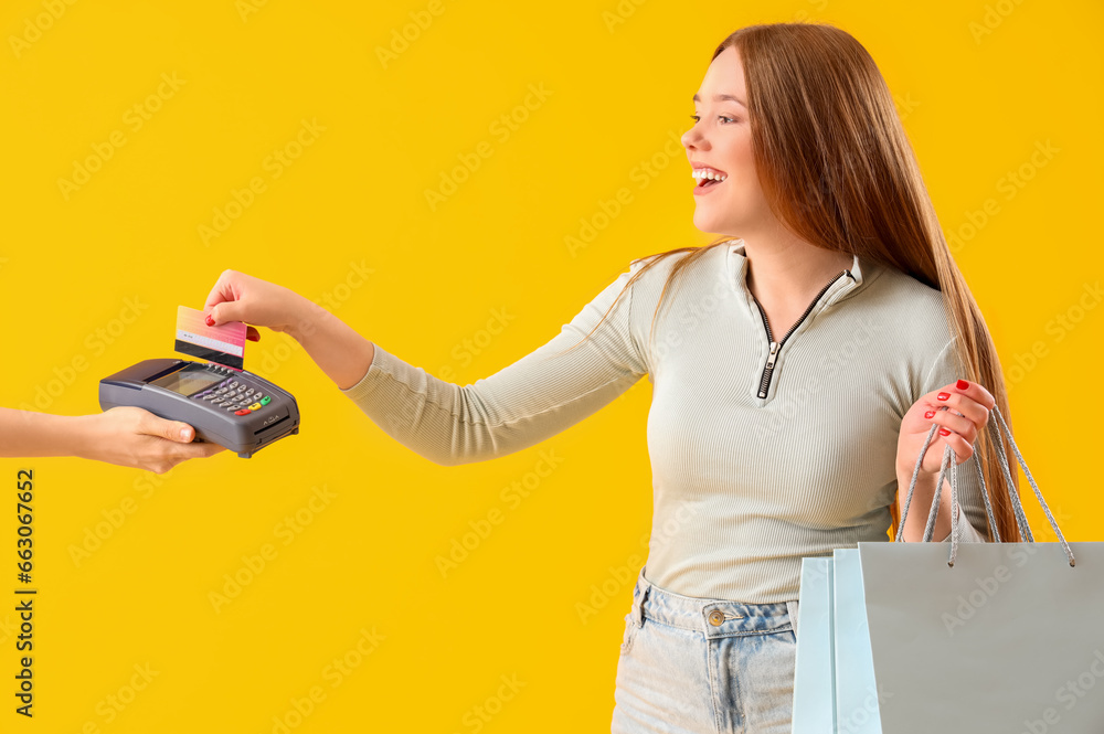 Φωτογραφία Young woman with credit card and shopping bags paying via terminal on yellow bac