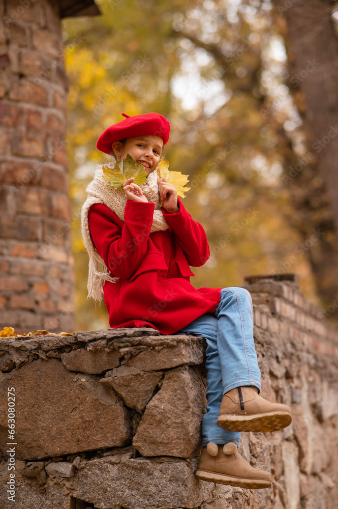 Smiling caucasian girl in a red coat and beret sits on a brick wall on a walk in autumn. 