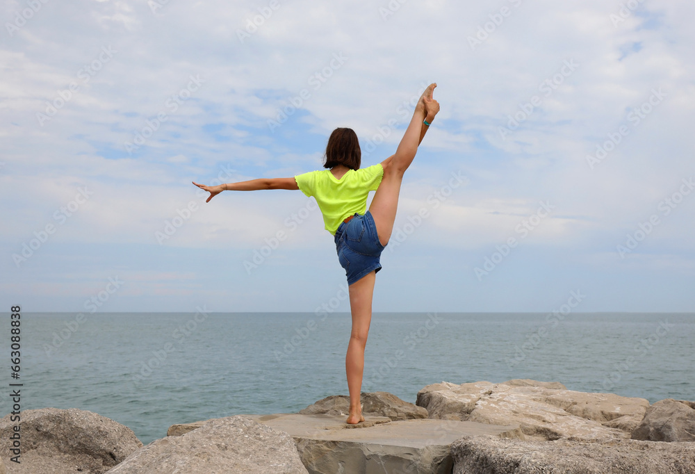 young girl performs gymnastic exercises on the rocks by the sea in jeans shorts in summer young girl performs gymnastic exercises on the rocks by the sea in jeans shorts in summer