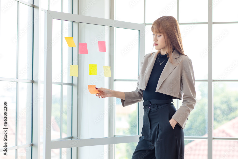 Asian professional successful young female businesswoman creative graphic designer in casual fashionable suit outfit standing sticking short note ideas on glass board in company office room