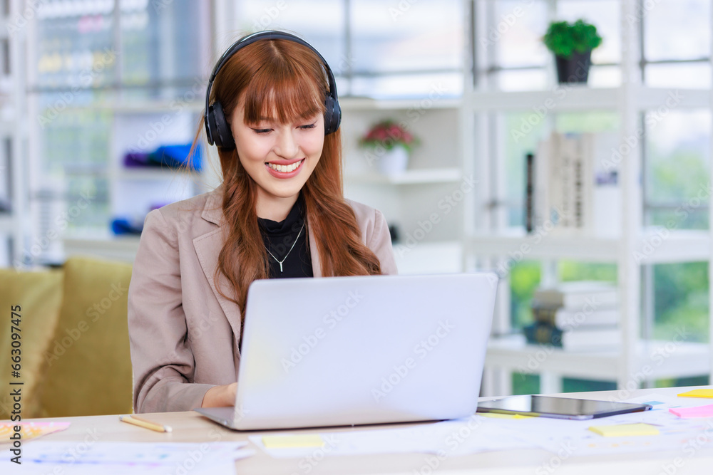 Asian professional successful young female businesswoman creative graphic designer in casual outfit sitting smiling wearing headphone listening to music playlist while working typing laptop computer Asian professional successful young female businesswoman creative graphic designer in casual outfit sitting smiling wearing headphone listening to music playlist while working typing laptop computer