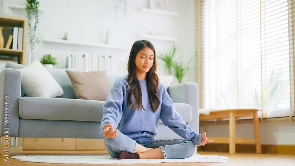 Happy young Asian woman practicing yoga and meditation at home sitting on floor in living room in lotus position and relaxing with closed eyes. Mindful meditation and wellbeing concept Happy young Asian woman practicing yoga and meditation at home sitting on floor in living room in lotus position and relaxing with closed eyes. Mindful meditation and wellbeing concept