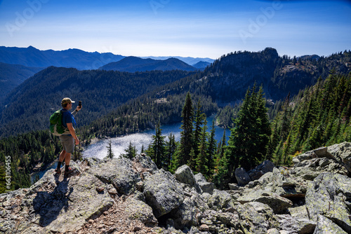 Adventurous athletic male hiker standing on top of a hill taking a photo of an alpine lake and a rugged mountain range on a beautiful sunny fall day in the Pacific Northwest. 