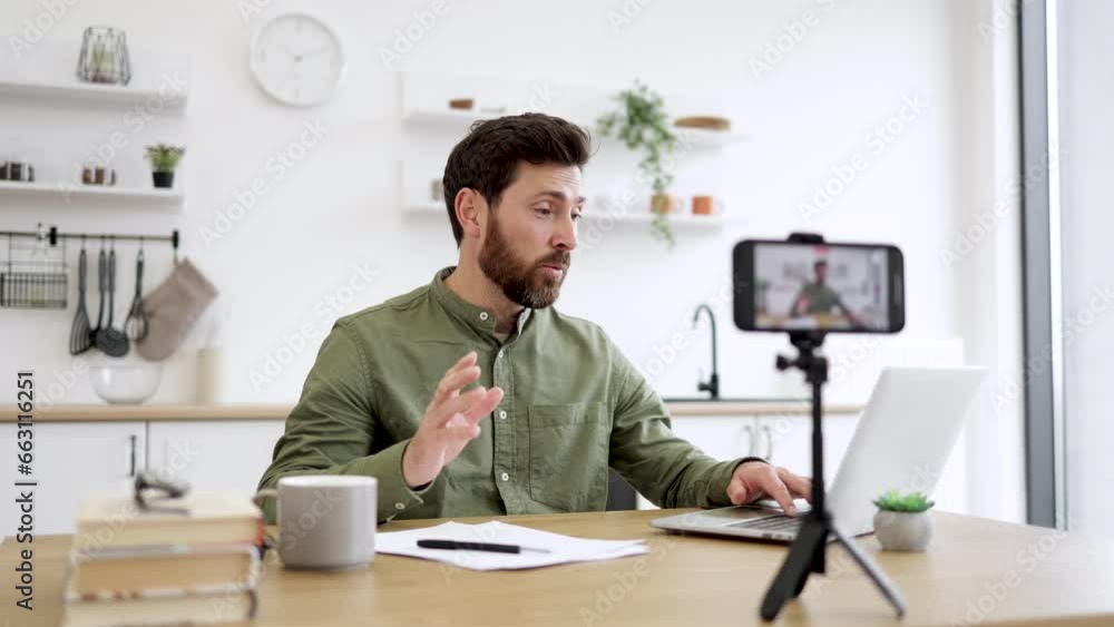 Modern smartphone fixed on tripod filming handsome caucasian man in casual wear working on portable laptop at home office. Blurred background. Technology, people and blogging concept. Modern smartphone fixed on tripod filming handsome caucasian man in casual wear working on portable laptop at home office. Blurred background. Technology, people and blogging concept.
