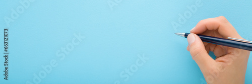 Young adult woman hand holding pen and writing on light blue background. Pastel color. Closeup. Empty place for text. Wide banner. Top down view.