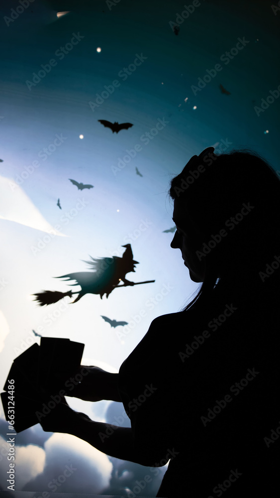Fototapeta premium the black shadow of a girl in profile with Tarot cards in hand cards in the background of clouds flying on a broomstick witch and bats