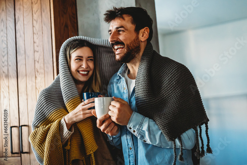 Obraz na plátně Happy young couple with hot drinks cuddling under warm blanket at home