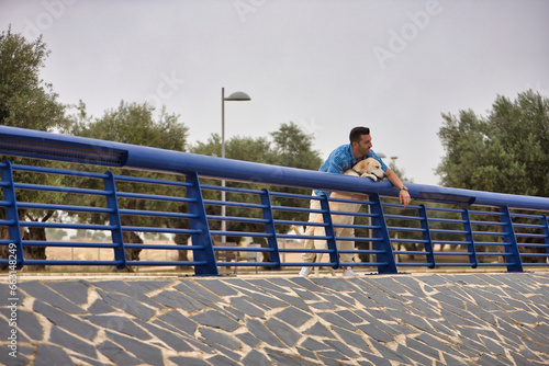 Young Hispanic man, leaning on a railing next to his dog looking both in the same direction. Concept, dogs, pets, animals, friends.