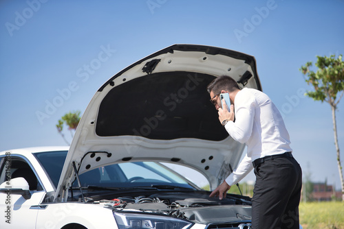 Young hispanic man, wearing a white shirt, calling the tow truck with his mobile, after suffering a breakdown in his white luxury car. Concept, cars, breakdowns, roadside assistance, crane. photo