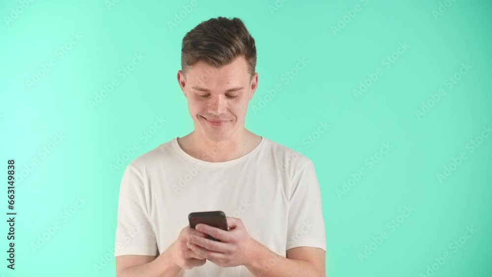 Young man is holding smartphone, using mobile apps, watching funny video in social media. Smiling man having fun chatting. Male has conversation with girlfriend on phone on blue background in studio. Young man is holding smartphone, using mobile apps, watching funny video in social media. Smiling man having fun chatting. Male has conversation with girlfriend on phone on blue background in studio.