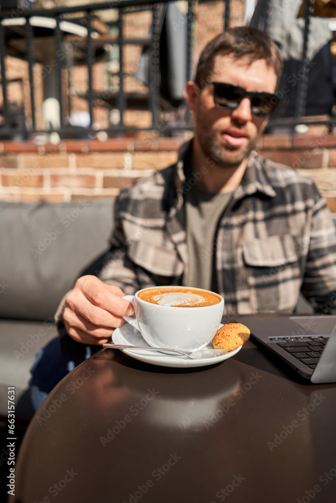 A man drinks coffee, holds a cup of hot Peanut latte in his hand. The table in the coworking space of the coffee shop. A cup of coffee on the veranda on a sunny day. Coffee break at the cafe. Close-up A man drinks coffee, holds a cup of hot Peanut latte in his hand. The table in the coworking space of the coffee shop. A cup of coffee on the veranda on a sunny day. Coffee break at the cafe. Close-up