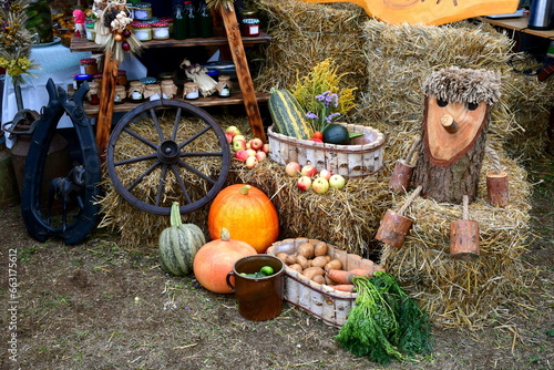 A close up on a set of decorations seen during a folk fair, including hay bales, autumn fruits, vegetables, crops, wooden figurines, rural equipment, and farming produce displayed for visitors